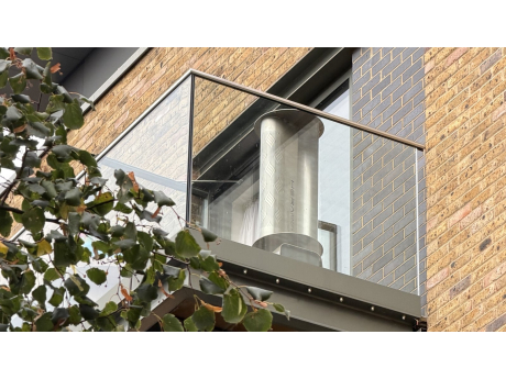 Una turbina eolica TESUP Hera Wind Pro dietro la balaustra in vetro di un balcone di un condominio in mattoni, con la vernice ancora come nuova dopo cinque anni all'aperto
