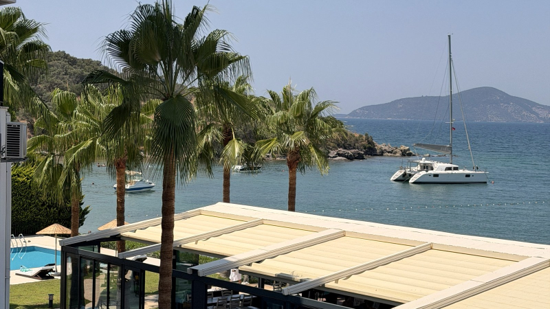 Med-Inn restaurant terrace and palm trees above Güllük Bay, with a catamaran at anchor and an island on the horizon