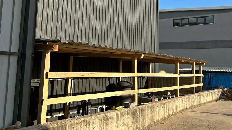 New timber-framed shelter with a corrugated roof along the outside wall of a TESUP production hall, housing the air compressor