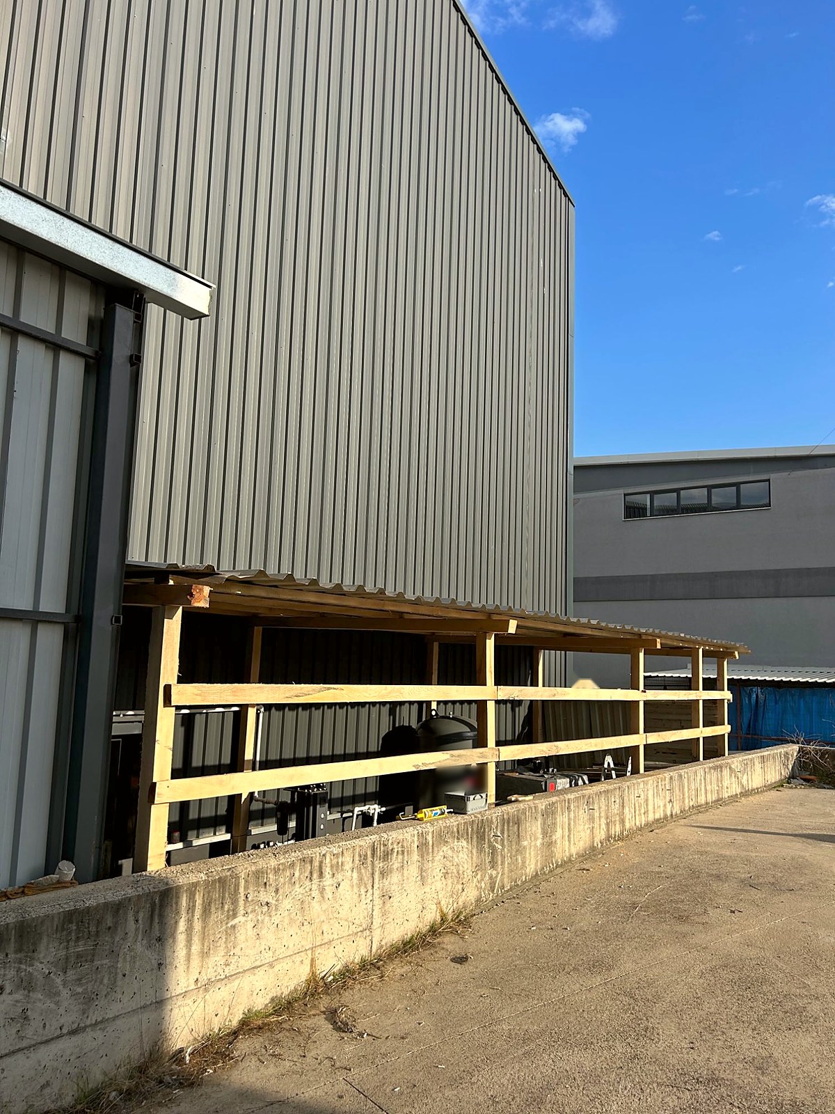 New timber-framed shelter with a corrugated roof along the outside wall of a TESUP production hall, housing the air compressor