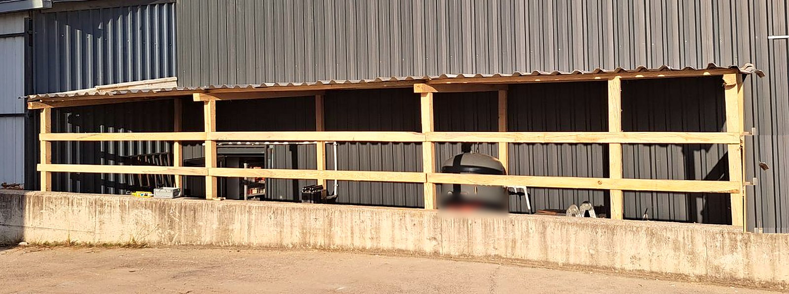 Full length of the new compressor shelter: timber posts and rails, corrugated roof and a concrete kerb along the dark grey wall of the production hall
