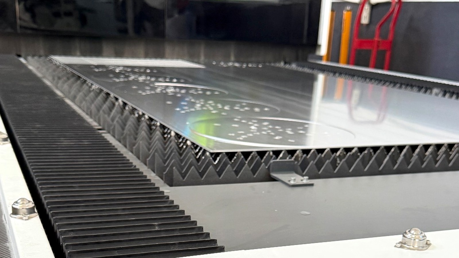 An aluminium sheet on the slat bed of a fibre laser cutter, a long curved blade profile cut through it, spatter beads across the surface