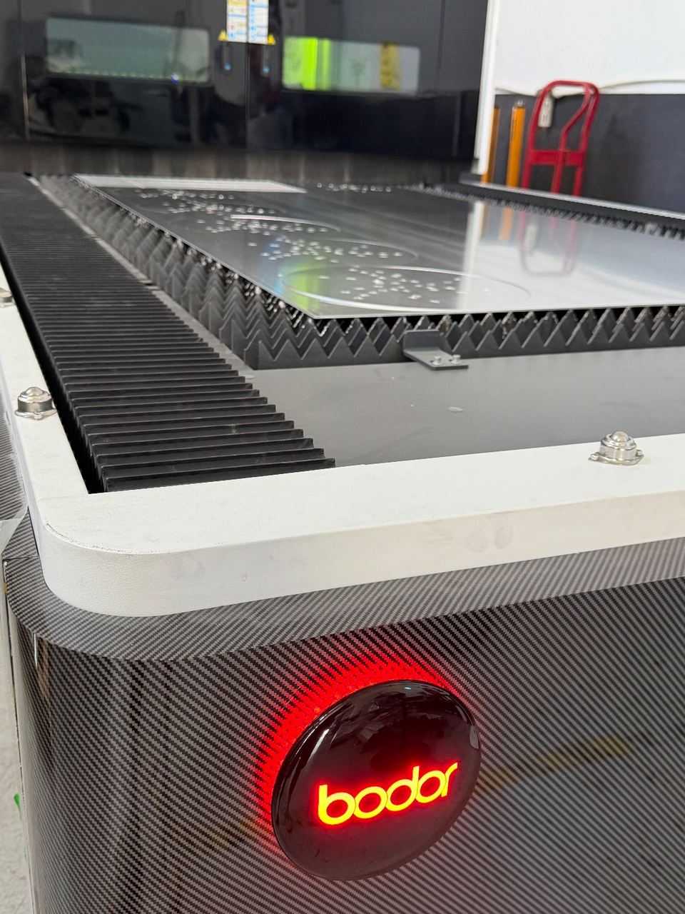 An aluminium sheet on the slat bed of a fibre laser cutter, a long curved blade profile cut through it, spatter beads across the surface