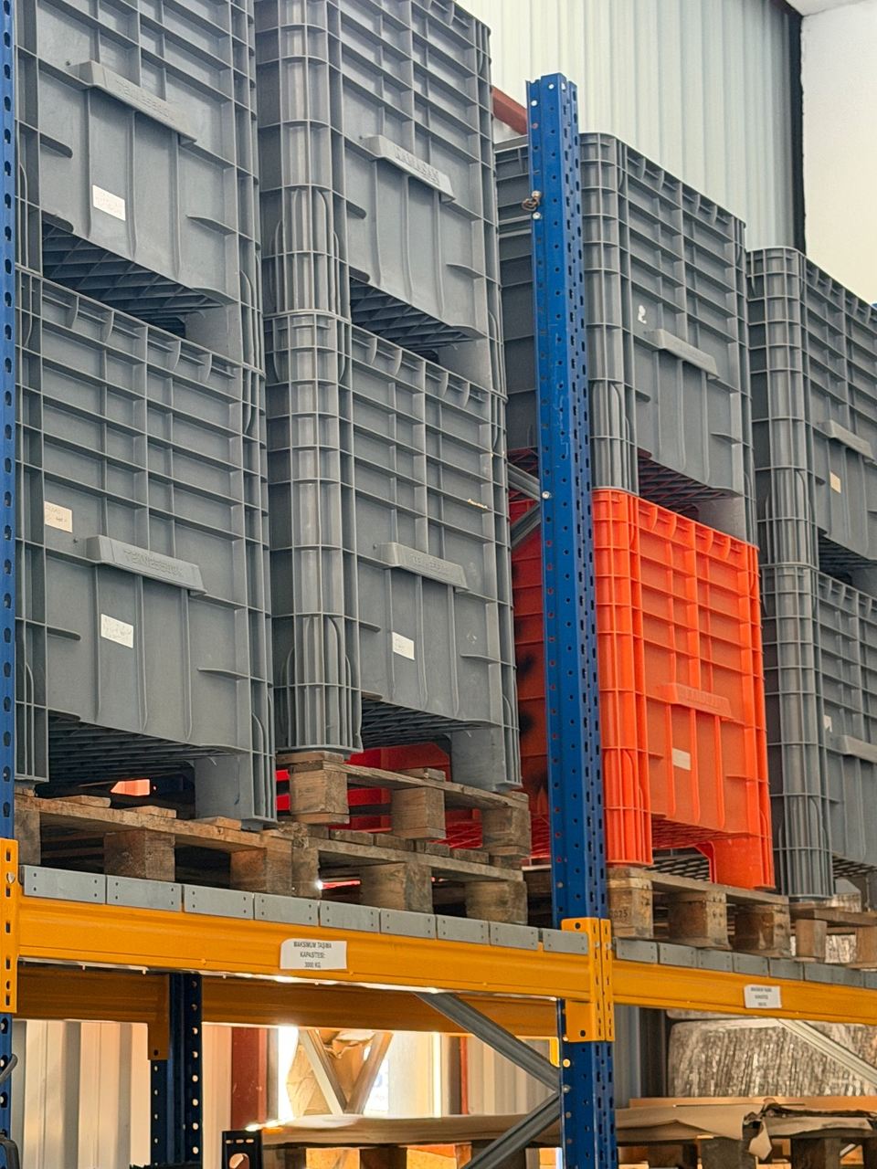 Warehouse pallet racking stacked with grey and orange returnable crates on wooden pallets, load-limit labels on the yellow beams