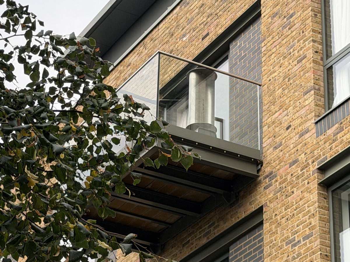 Une éolienne TESUP Hera Wind Pro sur le balcon vitré d'un appartement, vue depuis la rue à travers les branches d'un arbre