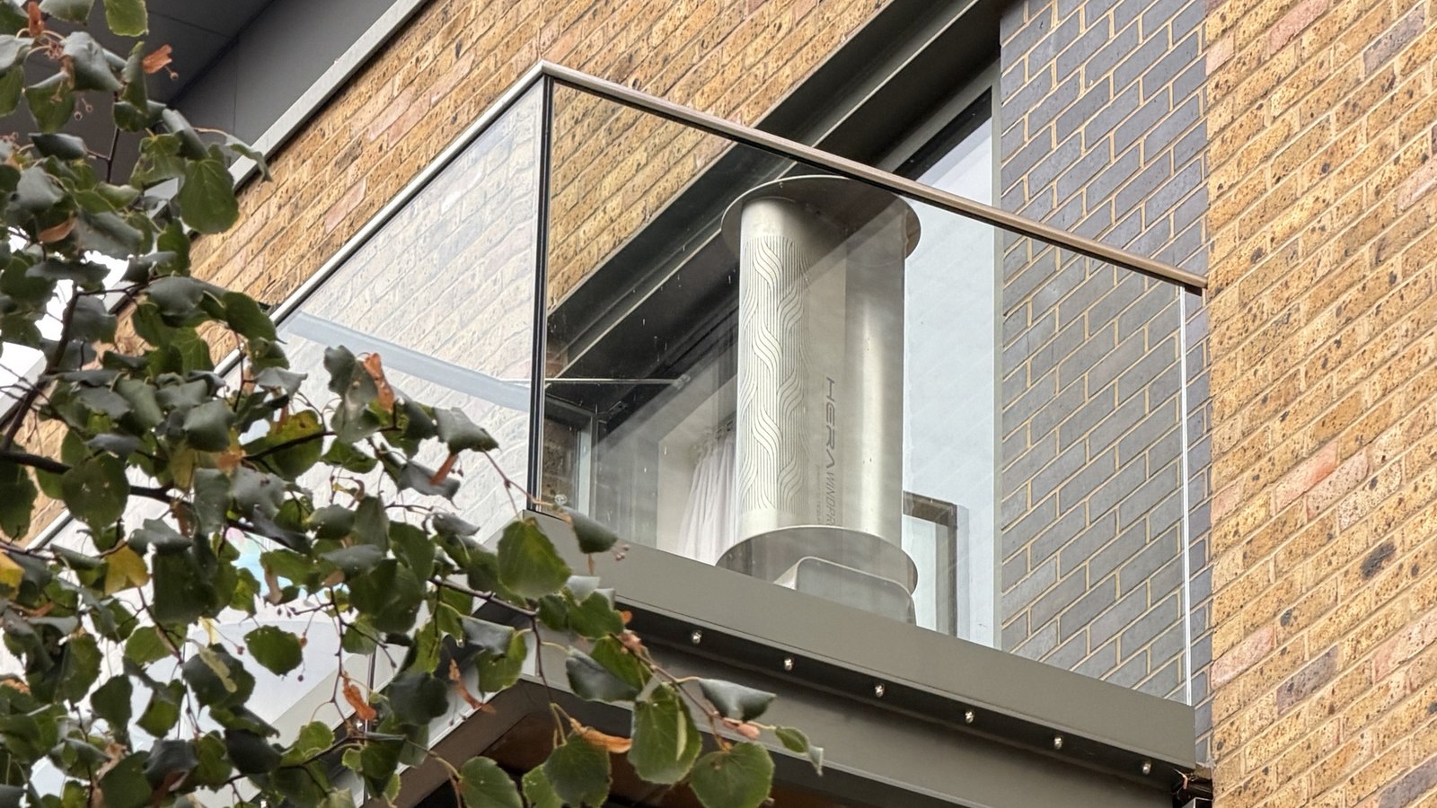 Une éolienne TESUP Hera Wind Pro derrière la balustrade en verre d'un balcon d'immeuble en brique, sa peinture toujours comme neuve après cinq ans en plein air
