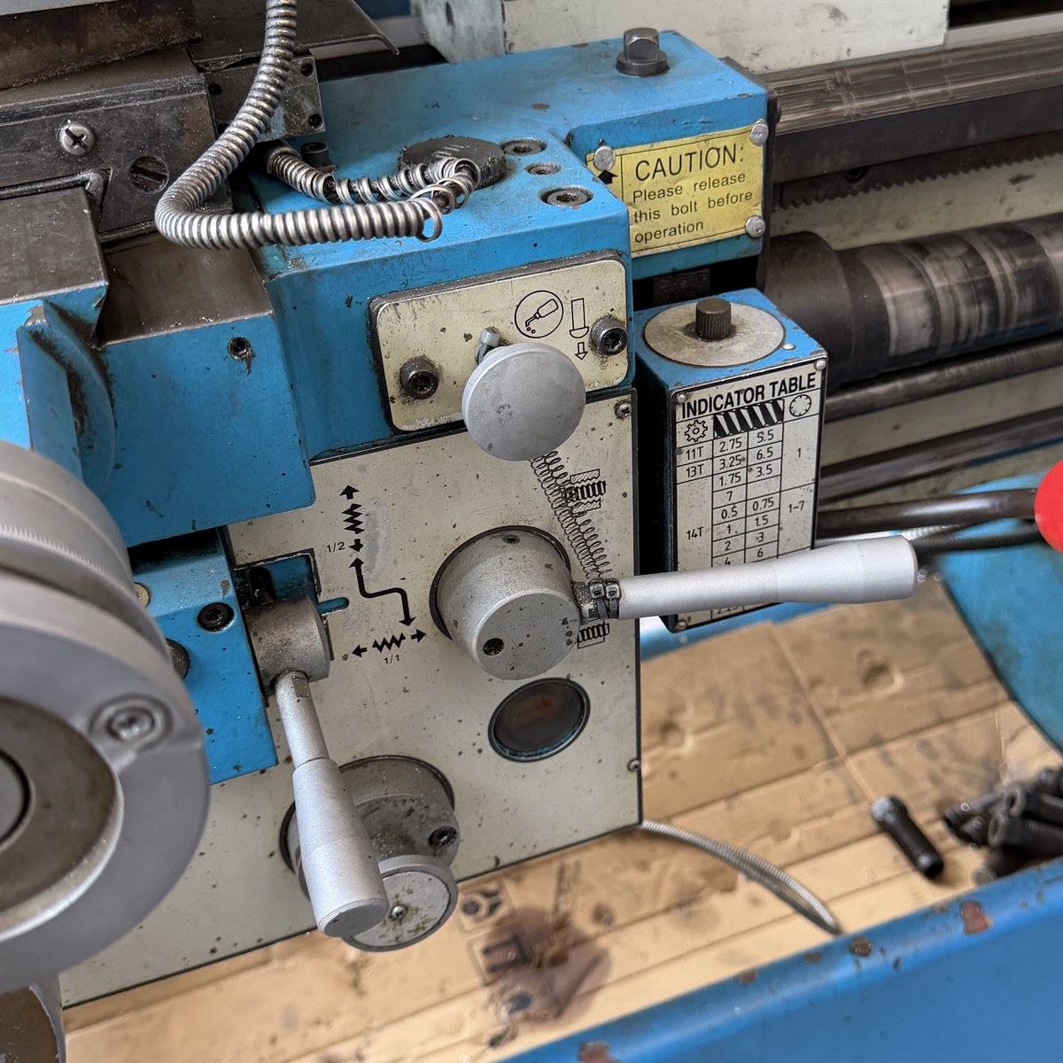 The carriage and apron of the lathe with the threading indicator dial, a yellow CAUTION sticker reading please release this bolt before operation, the half-nut lever with a red ball handle, the leadscrew and feed rod, and the cross slide handwheel in the foreground