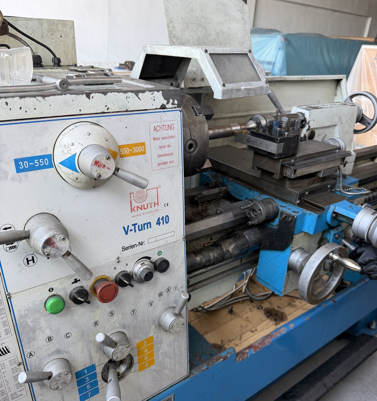 A KNUTH V-Turn 410 manual lathe in a workshop: headstock and control panel on the left with speed range labels, a chuck, a four-way tool post on the cross slide, the tailstock and the bed running to the right, with swarf in the chip tray