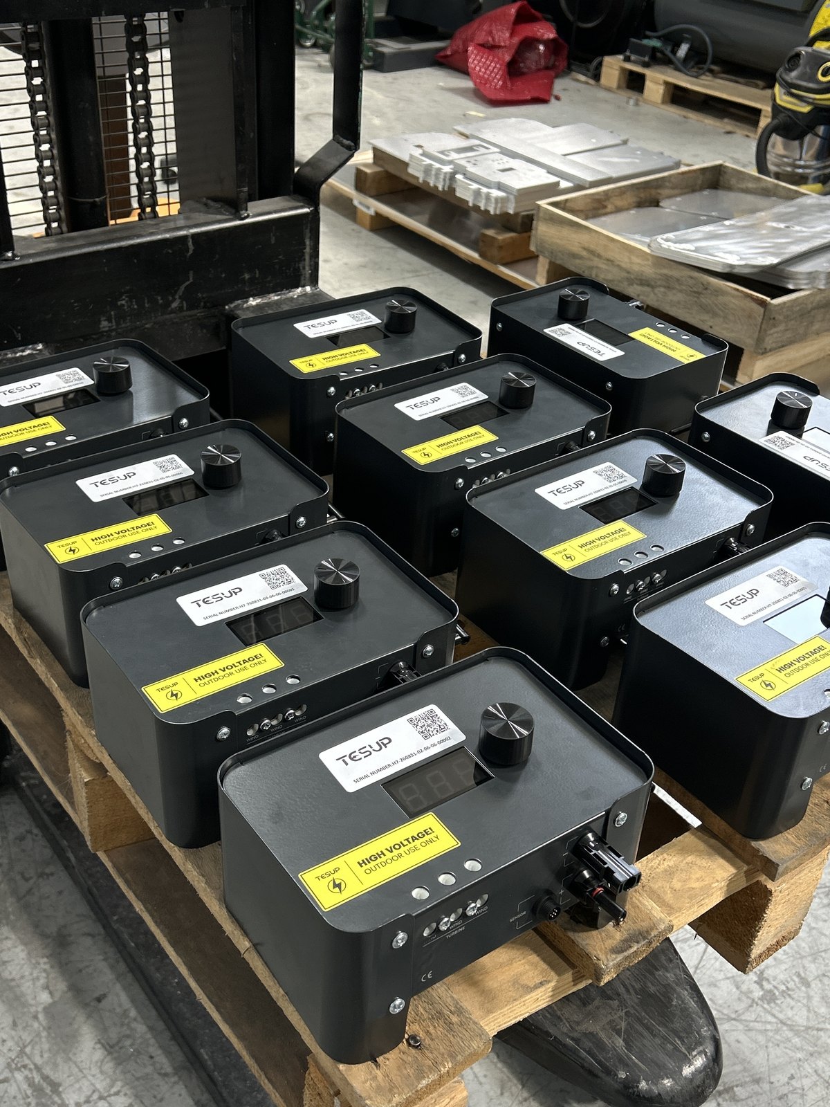 A batch of TESUP Magnum charge controllers arranged on a wooden pallet on the workshop floor, with a pallet stacker to the left and a pallet of flat laser-cut sheet metal blanks behind them