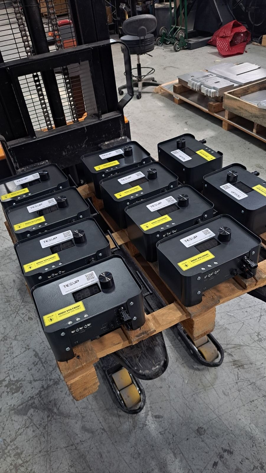 Ten TESUP Magnum charge controllers arranged in three columns on a wooden pallet, seen at an angle, with a pallet stacker and workshop equipment behind