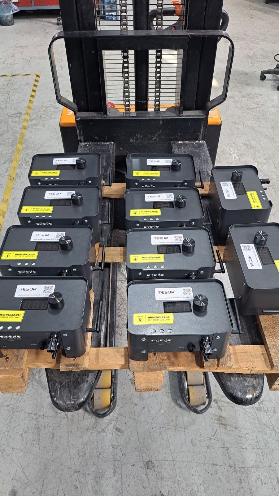 The same pallet of ten TESUP Magnum charge controllers seen from above on the forks of a pallet stacker, painted floor markings visible around it