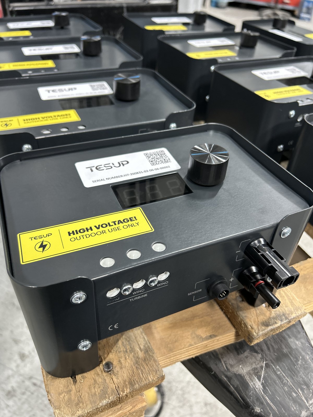 Close-up of the front panel of a TESUP Magnum charge controller on a wooden pallet: a TESUP serial label with a QR code, a three-digit display, a machined rotary knob, a yellow HIGH VOLTAGE OUTDOOR USE ONLY sticker, three indicators, three terminals labelled WIND under a TURBINE bracket, a SENSOR connector, MC4 connectors and a CE mark