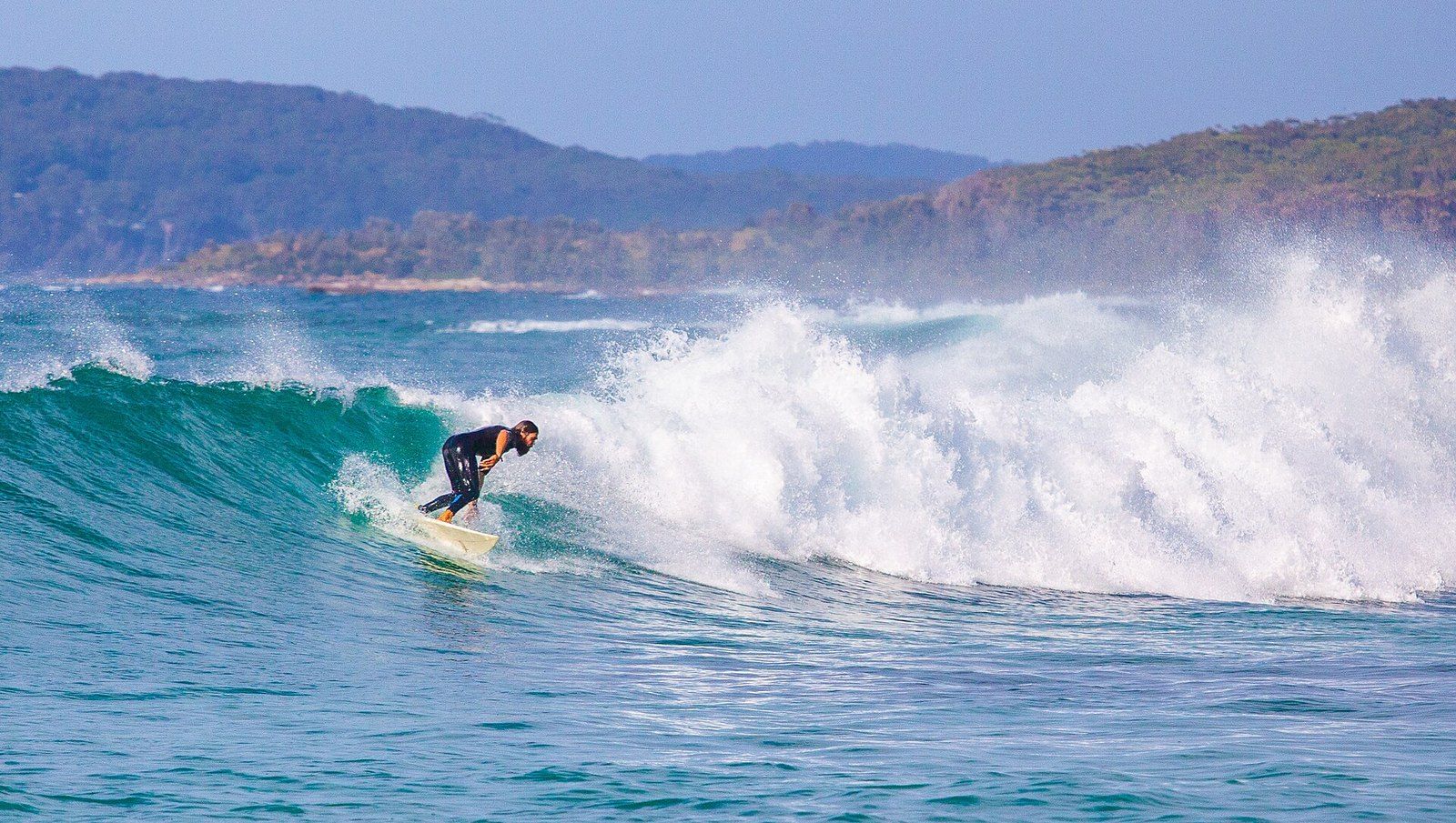 A surfer carving across a clean turquoise wave on the New South Wales coast, with bush-covered headlands behind