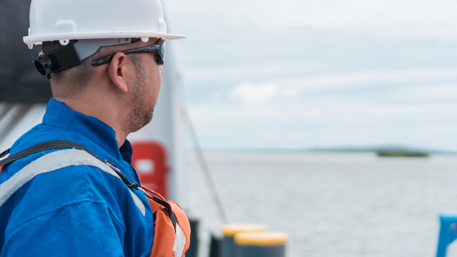 A deck officer in a white hard hat and blue coveralls looking out to sea from a ship’s rail