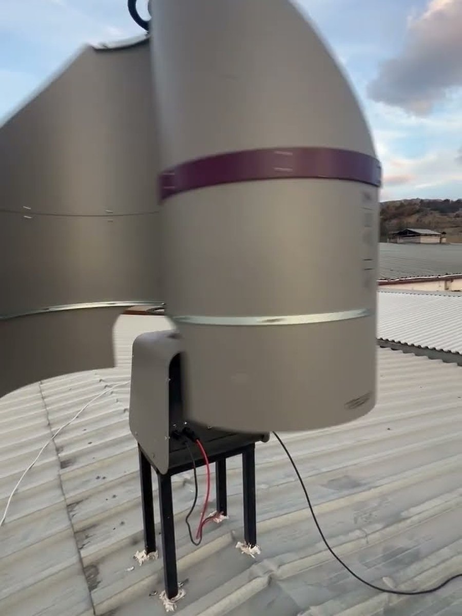 Uma turbina eólica vertical TESUP Atlas com pás cinzentas de alumínio a rodar num telhado de chapa ondulada sob um céu azul