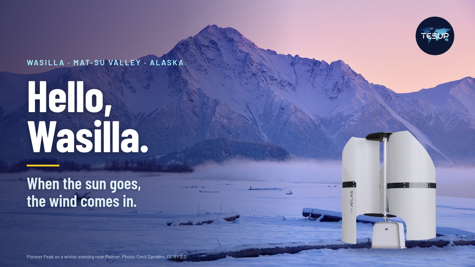 An Atlas wind turbine standing in the snow below Pioneer Peak near Palmer, Alaska, on a winter evening, beside the words Hello, Wasilla