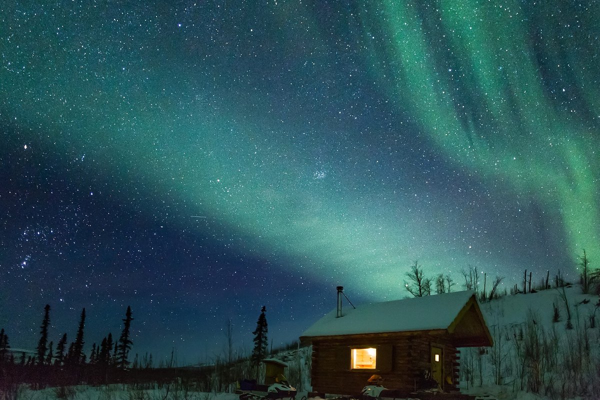 Green northern lights over a small log cabin in the snow, with a lit window, in Alaska